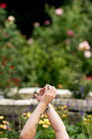 Close-up of two tattooed hands intertwined and raised against a colorful blurred garden background.