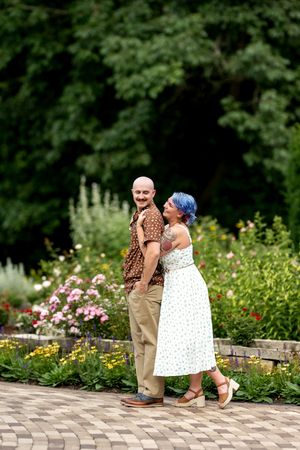 Couple posing on a brick path in a vibrant flower garden, woman with blue hair kissing mans cheek.