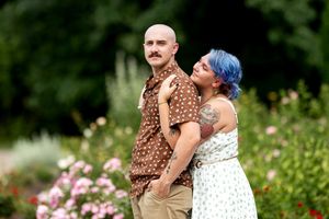 Woman with blue hair embracing a bald man with a mustache in a colorful blooming rose garden.