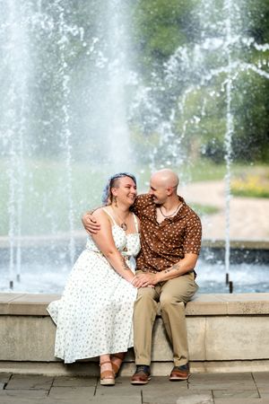 Couple sitting together smiling by a fountain in a park, woman in floral dress and man in brown polka dot shirt.