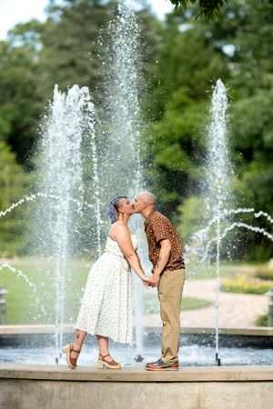 Couple kissing in front of a fountain, woman in white floral dress and man in brown shirt holding hands.