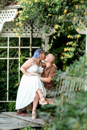Couple sharing kiss on garden bench under white lattice arbor with flowers.