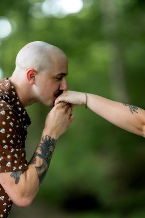 Tattooed bald man kissing a woman's hand outdoors with green trees in the background.