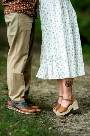 Close-up of couple's feet, man in brown leather shoes and woman in wedge sandals with ankle tattoo.