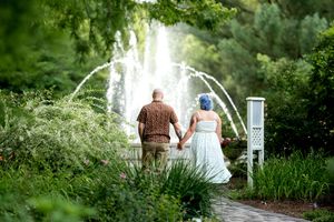 Couple holding hands while gazing at a beautiful garden fountain surrounded by lush green foliage.