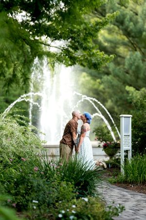 Couple sharing a romantic kiss in a lush garden with an elegant fountain and arching water jets behind them.