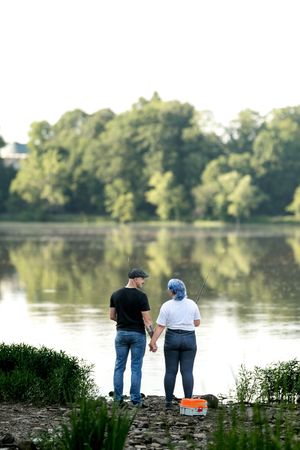 Couple fishing hand-in-hand by a calm reflective lake surrounded by green trees.
