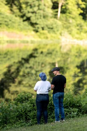 Couple standing together fishing at a peaceful lake with lush green tree reflections.