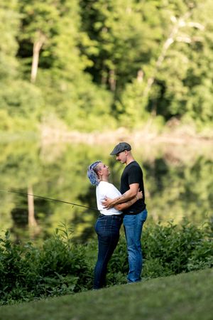 Couple embracing near a reflective lake, woman with blue hair in white top and man in black shirt.