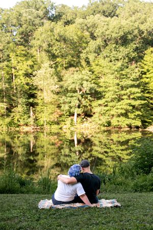 Couple sitting on a blanket by a serene lake, arms around each other enjoying the view.