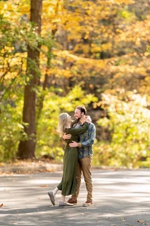 Happy couple dancing together on sunlit path surrounded by vibrant yellow autumn trees.