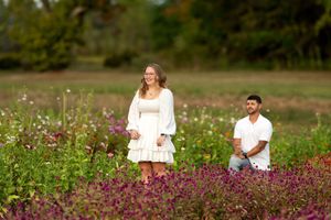 Man on one knee proposing to woman in white dress in a vibrant purple and pink wildflower meadow.