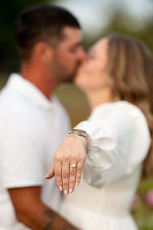 Engaged couple kissing while woman displays diamond engagement ring in sharp focus in the foreground.
