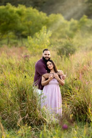 Couple embracing warmly in a wildflower meadow with golden sunrays streaming through trees behind them.