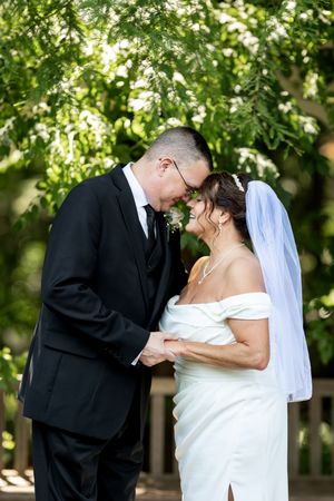 Bride and groom touching foreheads tenderly under green trees on wedding day.