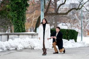 Man proposing on one knee to a surprised woman in a white coat in a snowy winter park setting.