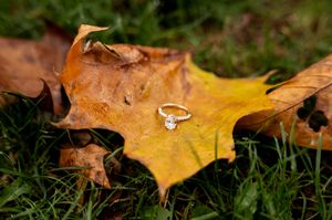 Diamond engagement ring resting on a golden autumn maple leaf surrounded by green grass.