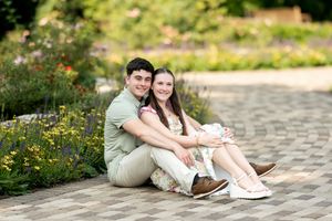 Happy engaged couple sitting together on garden path surrounded by summer flowers.