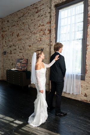 Bride in white lace gown taps grooms shoulder during first look in rustic brick venue.