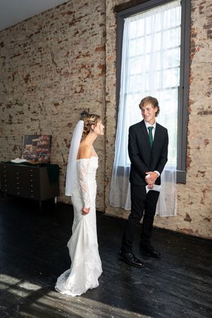 Groom smiles at bride during emotional first look in sunlit rustic brick room.