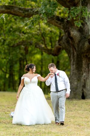 Groom kisses bride's hand as they walk together on grass beneath a large oak tree at their wedding.