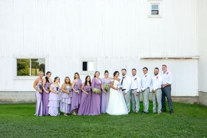 Wedding party poses in front of a white barn, bridesmaids in lavender dresses and groomsmen in gray suits.