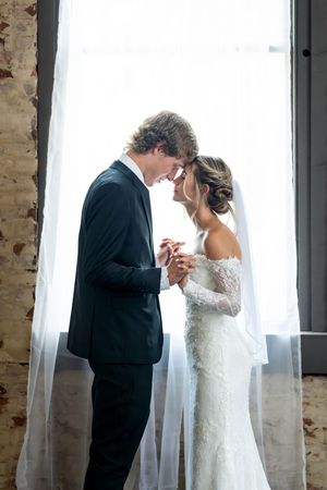 Bride and groom hold hands forehead to forehead in romantic backlit window portrait.