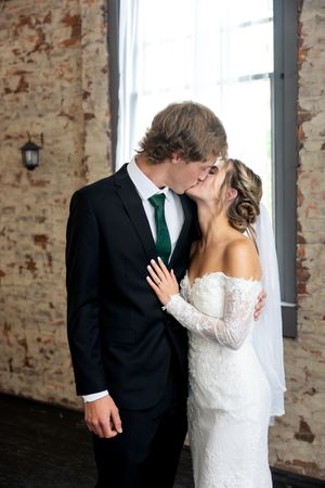 Bride and groom share a kiss in front of rustic exposed brick wall at venue.