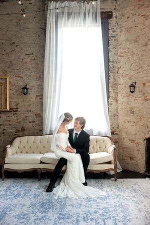 Bride and groom sit together on vintage tufted sofa in front of large bright window.