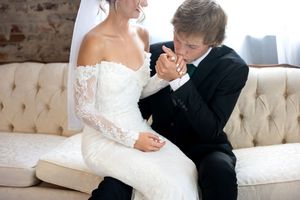 Groom kisses brides hand while seated together on cream tufted sofa at wedding venue.