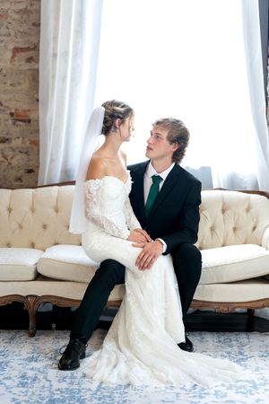 Bride and groom gaze at each other while seated on elegant tufted sofa at wedding.