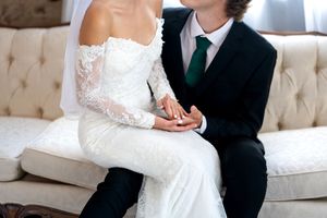 Bride and groom sitting together on elegant sofa, holding hands, bride in lace off-shoulder gown.