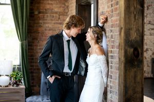 Bride in lace gown and groom in black suit share a tender moment by a rustic brick wall.