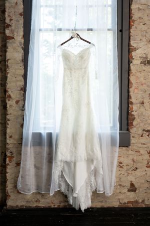 Lace mermaid wedding gown hanging on personalized hanger in window of rustic brick-walled venue.