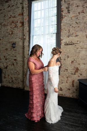 Mother helping bride button her lace wedding dress in a rustic brick-walled bridal suite before the ceremony.