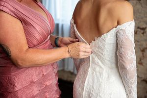 Woman in pink dress buttoning the back of a brides white lace wedding gown.