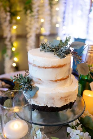 Elegant two-tier semi-naked wedding cake decorated with eucalyptus and greenery on a glass stand.