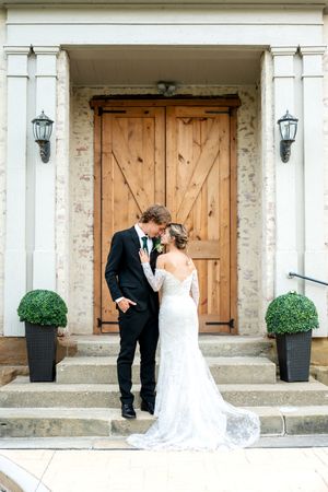 Bride and groom share a kiss on steps before rustic wooden doors of a charming wedding venue.