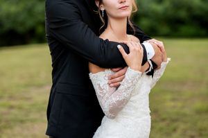 Groom tenderly embraces bride in lace off-shoulder wedding gown during outdoor portrait session.