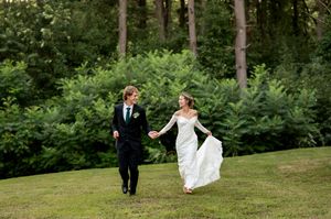 Bride and groom walk hand in hand across a lush green lawn surrounded by forest trees.
