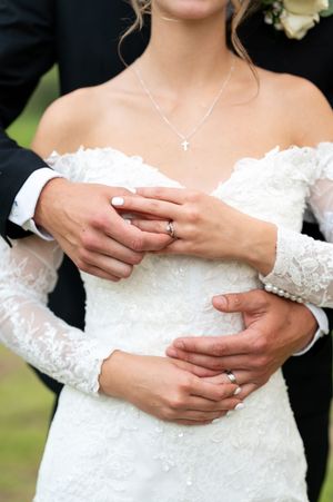 Close-up of bride and groom displaying wedding rings, bride wearing cross necklace and lace gown.