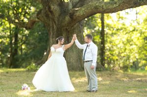 Bride and groom high-five under a large oak tree in a sunlit green field during outdoor wedding photos.
