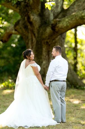 Bride in white gown and groom in gray suit hold hands while gazing at each other under a large tree.