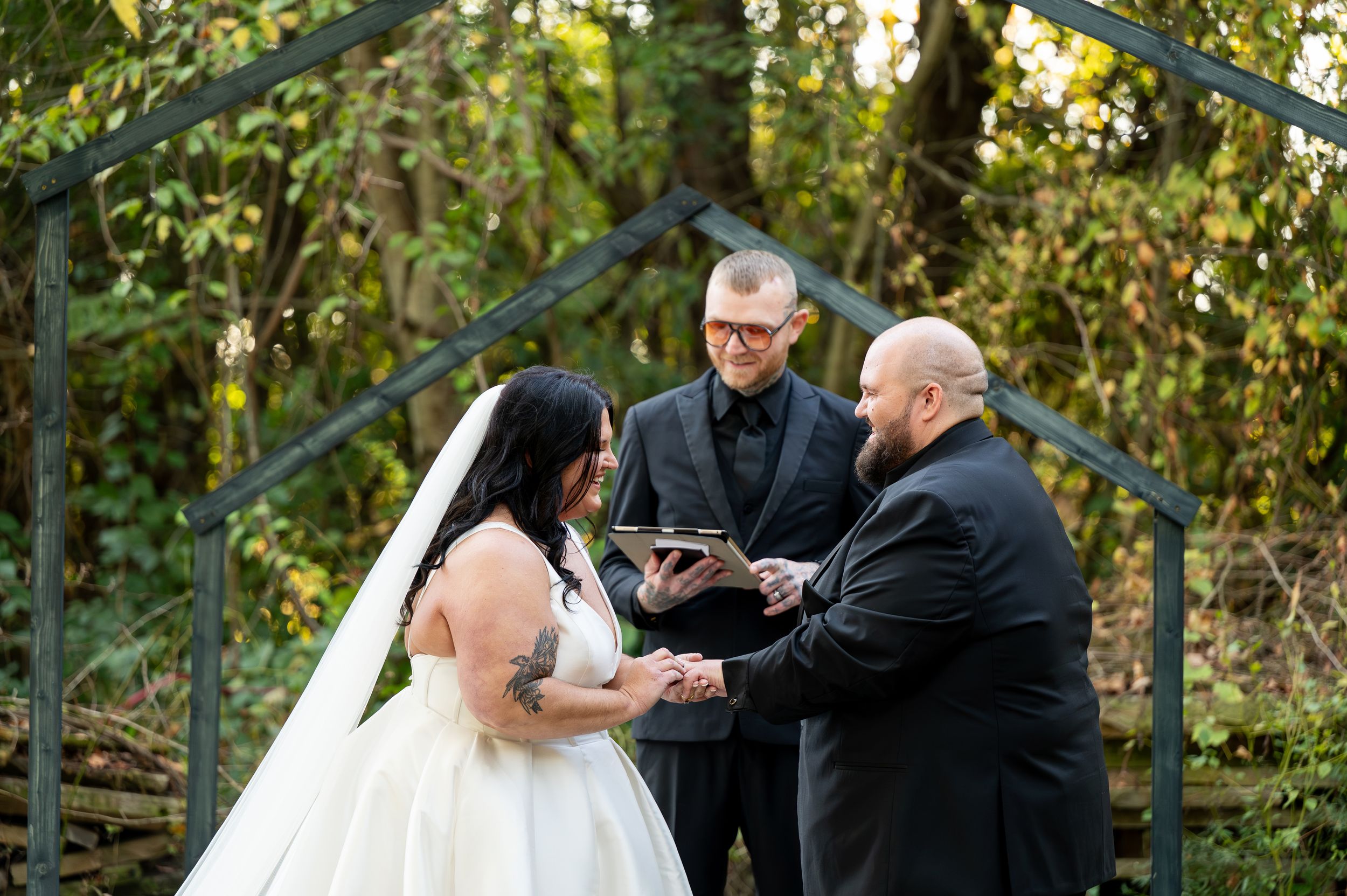 Bride and groom exchange vows with officiant at an outdoor woodland wedding ceremony beneath a rustic metal arch.