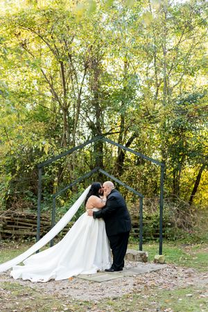 Newlyweds share their first kiss under a rustic metal arch surrounded by lush autumn forest greenery.