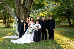 Wedding party poses in black attire with bride and groom before a floral arch beside a large tree outdoors.