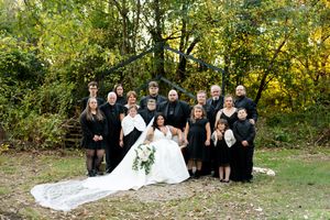 Large wedding party in black attire gathers with bride in white gown for a group portrait in autumn woods.
