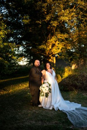 Bride holding white bouquet and groom pose together in warm golden evening light beneath a large oak tree.