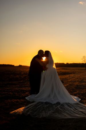 Silhouette of bride and groom sharing a romantic kiss against a stunning golden sunset over an open field.
