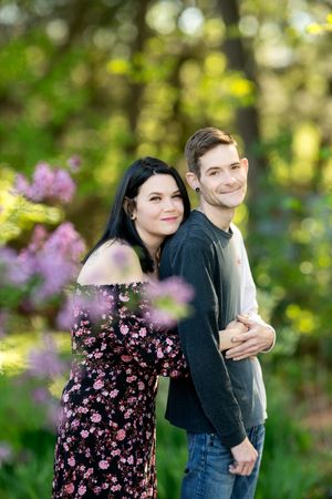 Couple embracing and smiling together outdoors surrounded by blooming pink flowers in a lush garden.
